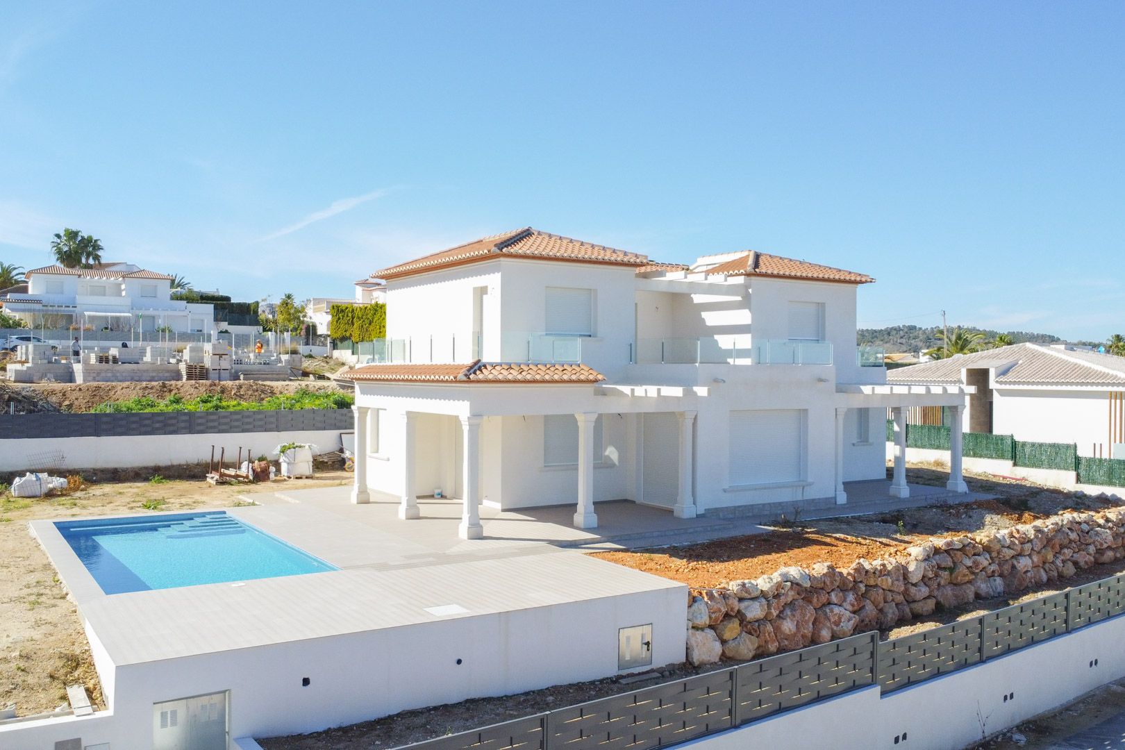 Vue extérieure de la villa blanche en fin de construction, présentant une piscine rectangulaire, des porches à colonnes et des terrasses avec garde-corps en verre.