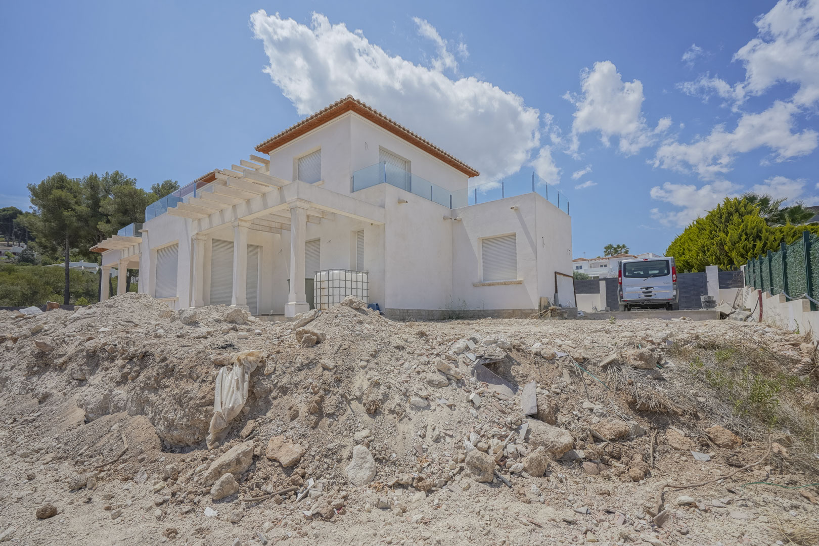 Vue extérieure de la villa en phase finale de construction, présentant des façades blanches, des pergolas à colonnes et des terrasses avec garde-corps en verre.