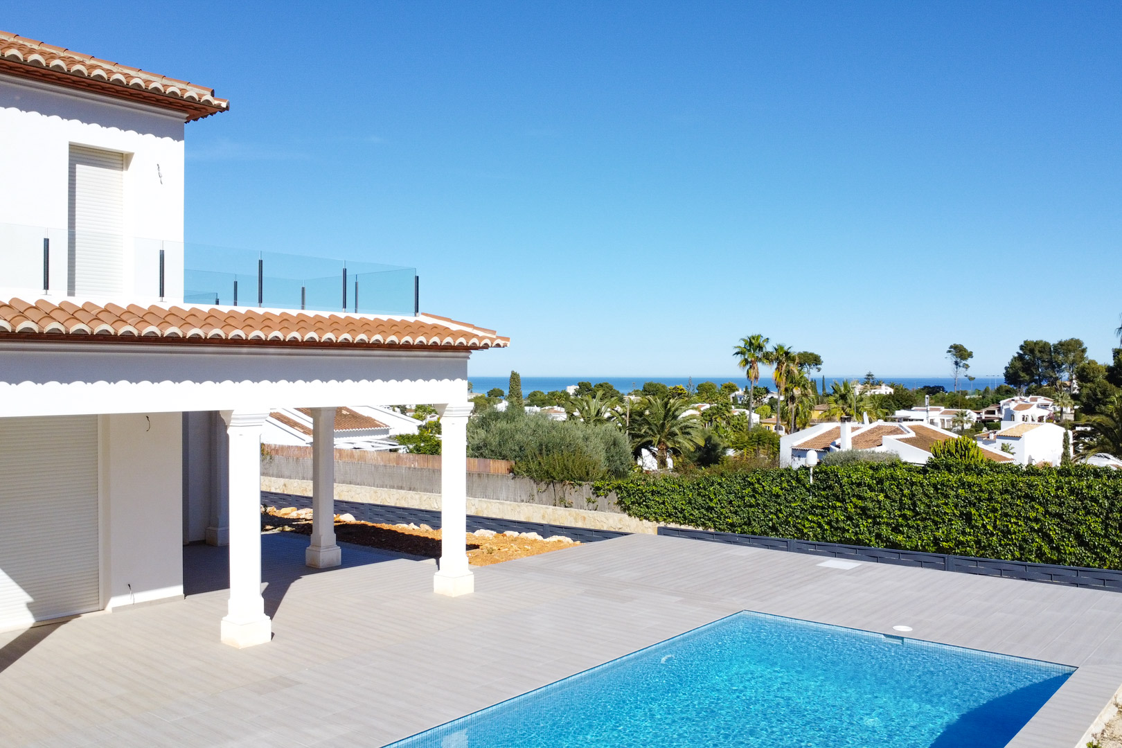 Vue extérieure montrant la terrasse en grès cérame, la piscine en mosaïque bleue et le porche à colonnes. L'arrière-plan offre une vue dégagée sur la mer Méditerranée.