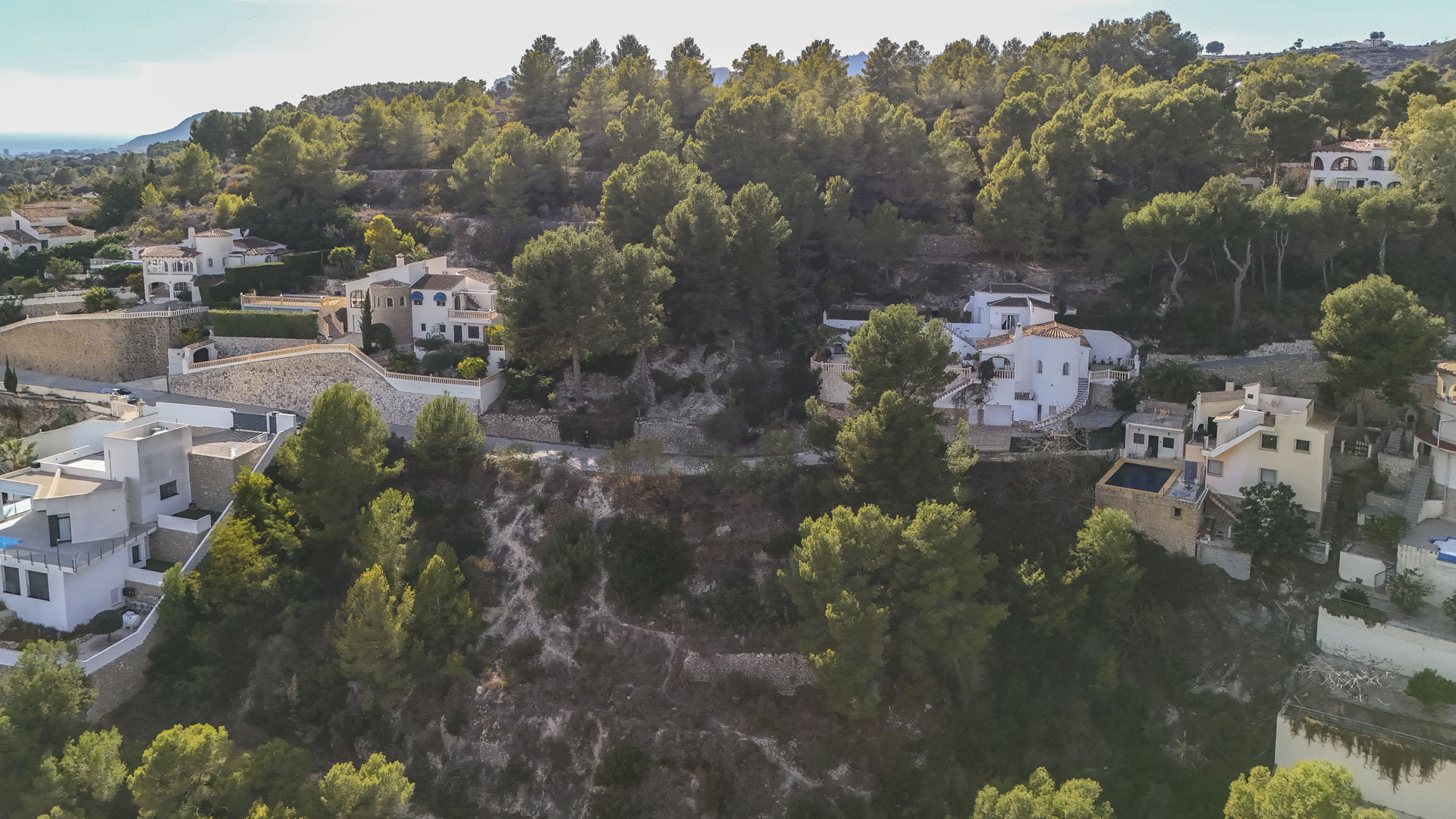 Vue aérienne de parcelles résidentielles à Benissa, nichées dans une pinède dense avec des murs en pierre et une vue lointaine sur la mer Méditerranée.