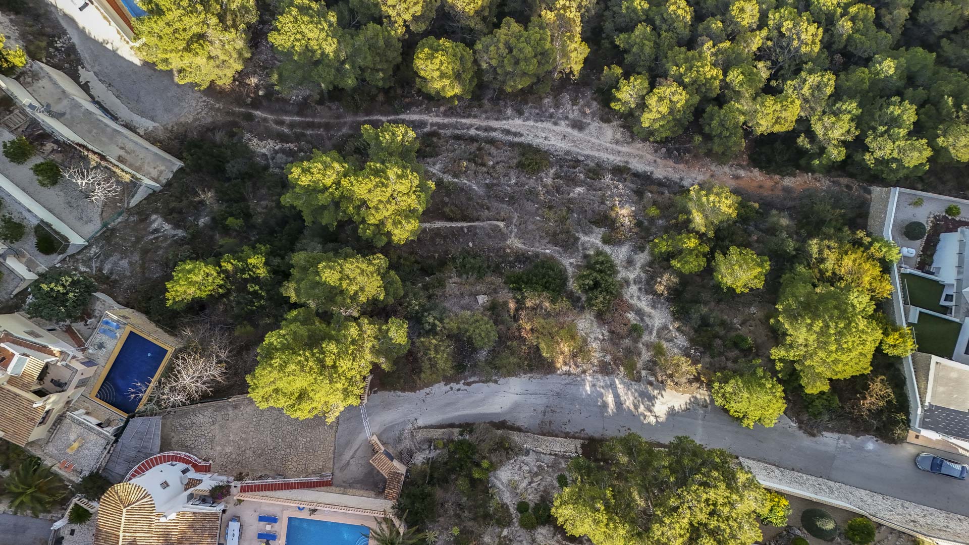 Vue aérienne zénithale du terrain urbain à Benissa, illustrant sa topographie en pente douce, sa végétation de pins et son accès par une route goudronnée.