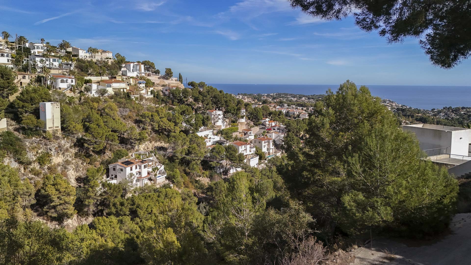Vue panoramique depuis le terrain surélevé vers la mer Méditerranée, avec des villas blanches nichées dans une colline boisée de pins sous un ciel dégagé.