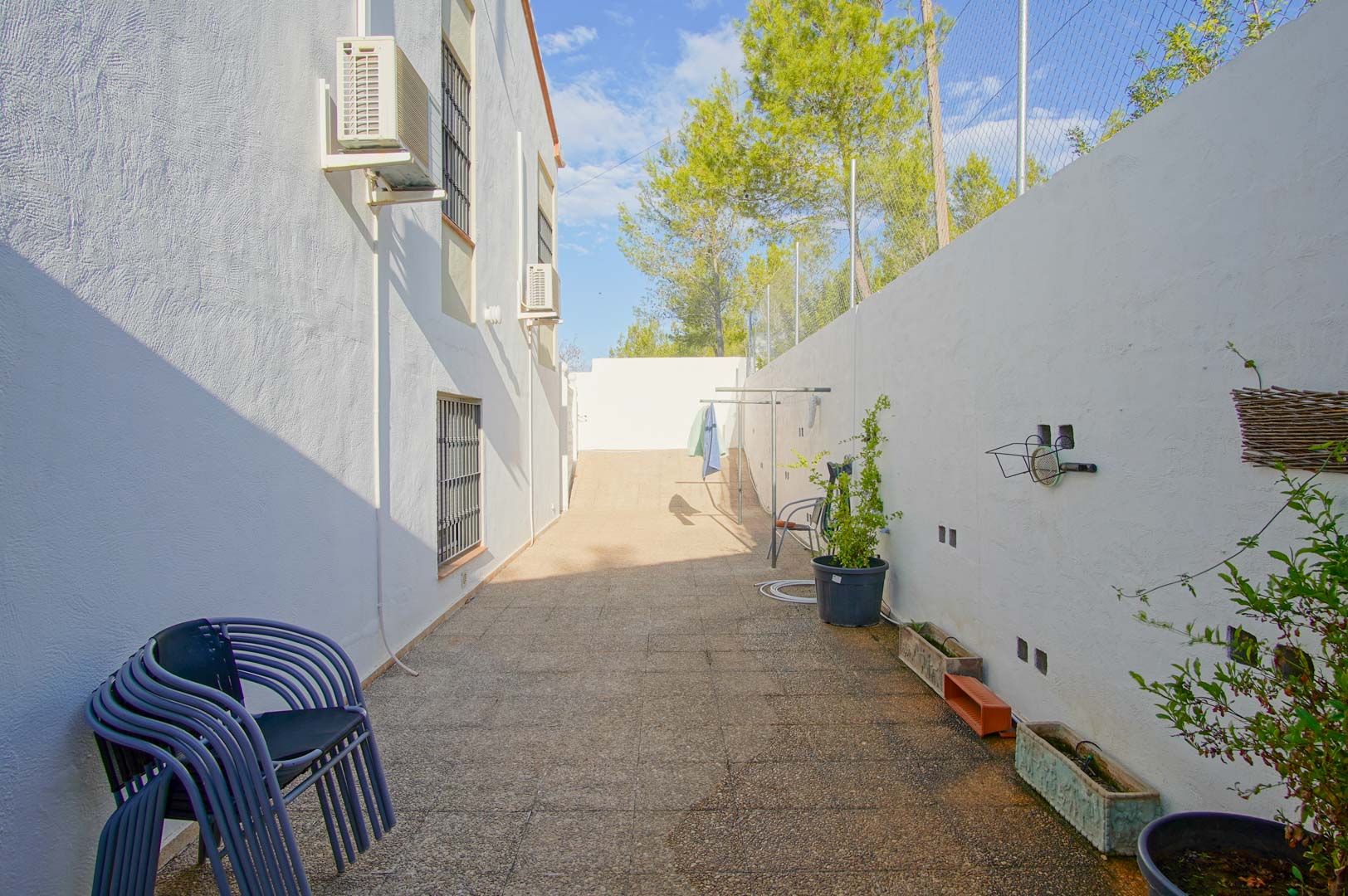 Allée latérale pavée avec murs blancs, unités de climatisation et clôture de sécurité, entourée de pins méditerranéens typiques de Las Rotas.