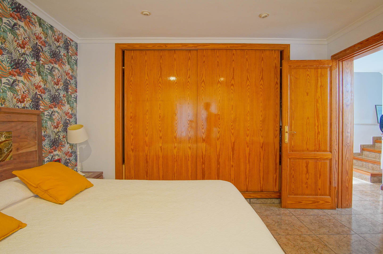 Chambre avec grand placard intégré en bois, mur d'accent au motif tropical et sol en carrelage. La porte ouverte mène vers un couloir avec escalier.