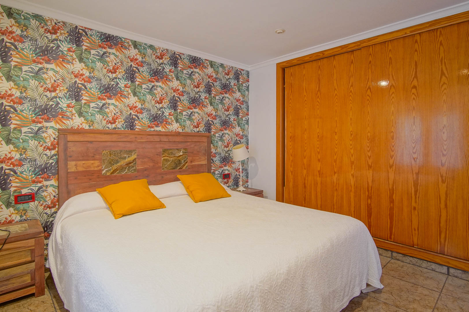 Chambre avec papier peint tropical, tête de lit en bois avec inserts en pierre et un grand placard intégré en bois naturel.