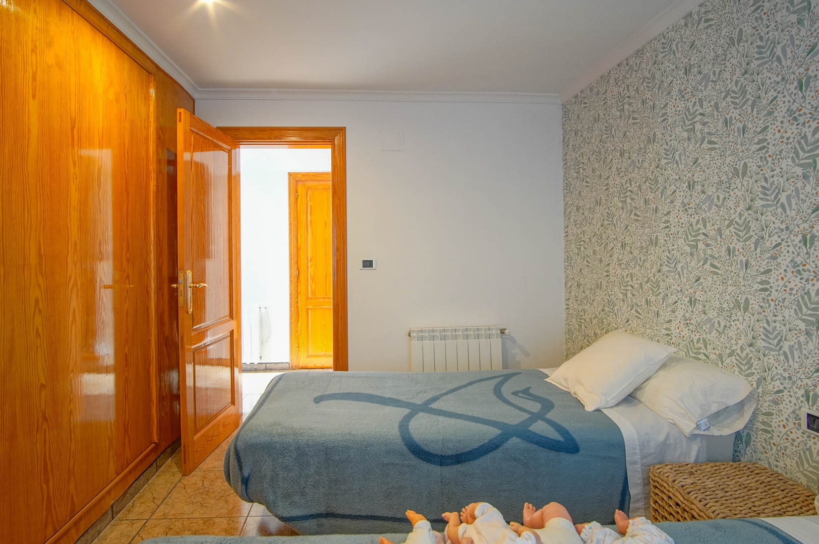 Chambre avec placard intégré en bois verni, papier peint aux motifs botaniques, sol en grès cérame et radiateur mural.