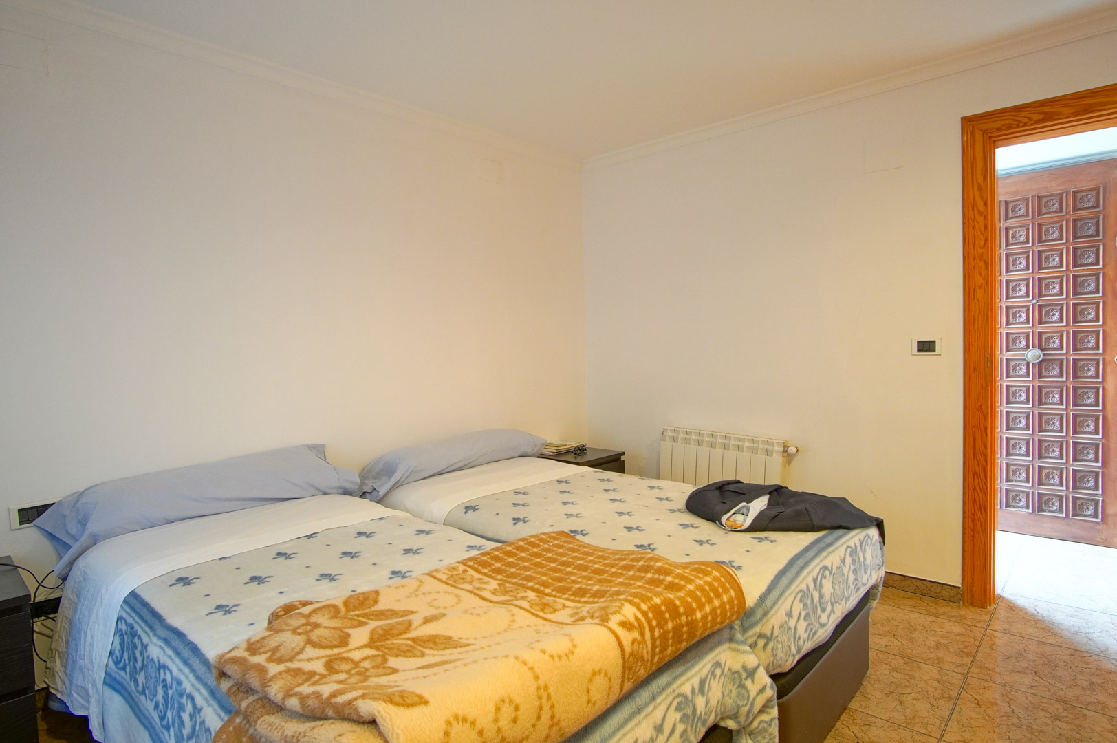 Chambre fonctionnelle avec lits jumeaux, murs blancs et sol en céramique. Elle comprend un radiateur et une porte en bois aux panneaux sculptés.