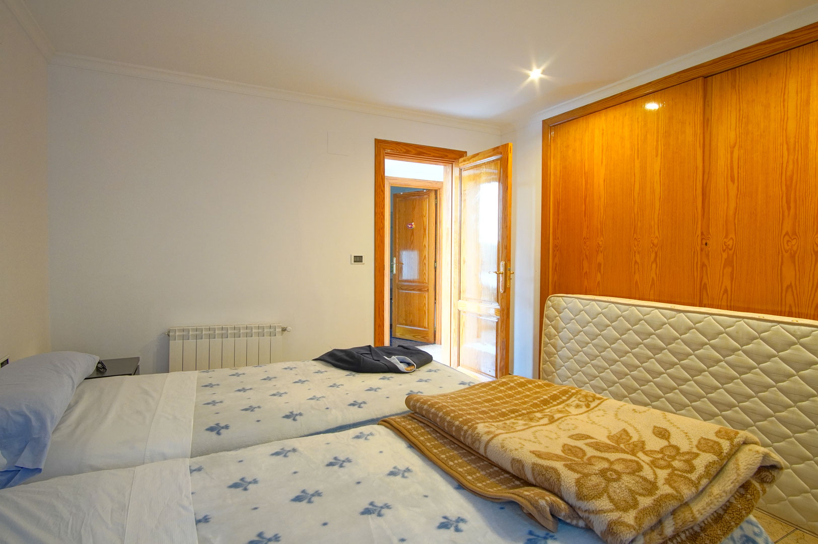 Chambre lumineuse avec de grands placards intégrés en bois, deux lits simples et un radiateur mural. Les finitions en bois assorties créent une atmosphère chaleureuse.