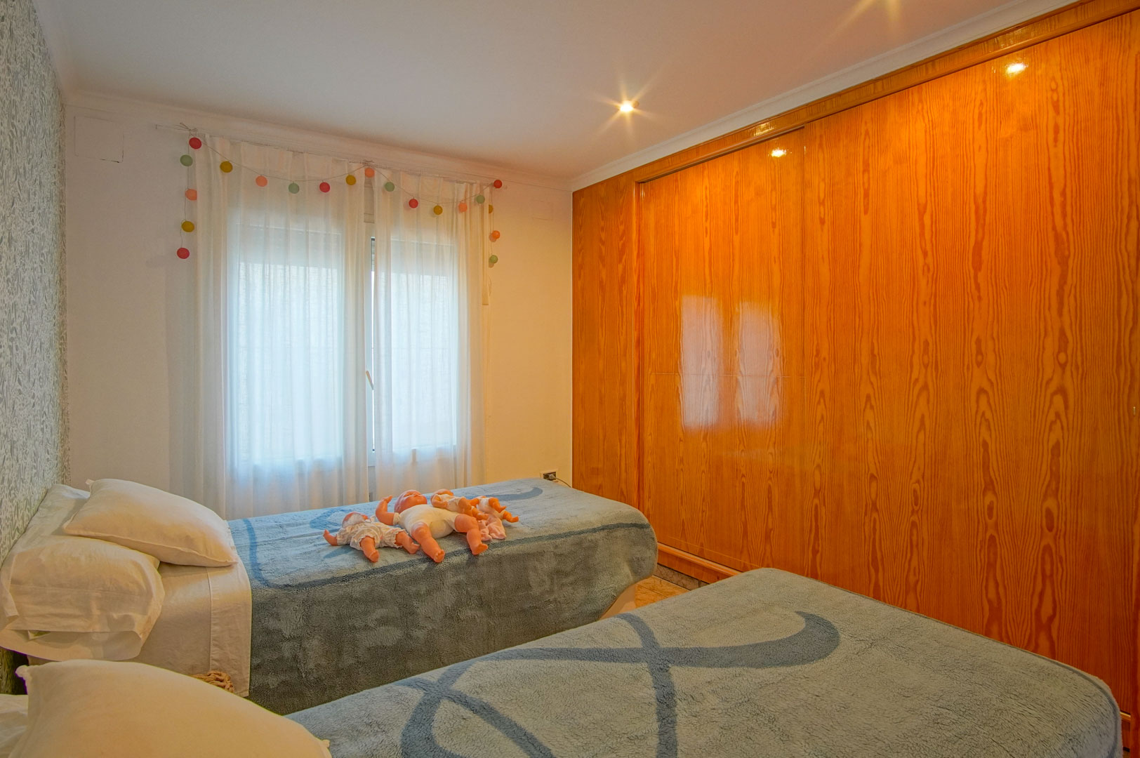 Chambre lumineuse avec deux lits simples, papier peint floral et un grand placard intégré en bois naturel aux finitions brillantes.