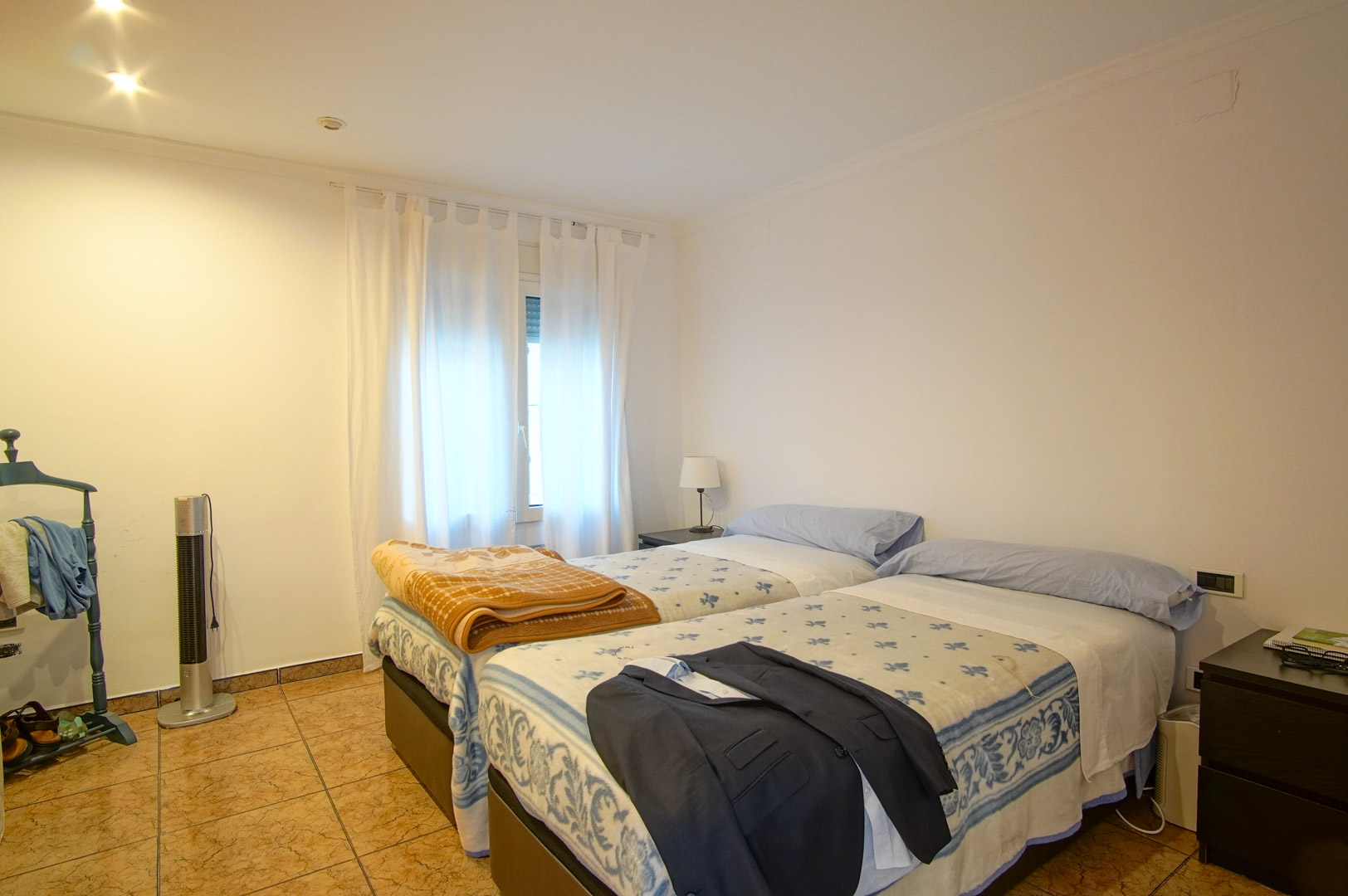 Chambre lumineuse avec deux lits simples, sol en grès cérame et murs blancs. Elle dispose d'un éclairage encastré et d'une fenêtre apportant une lumière naturelle douce.