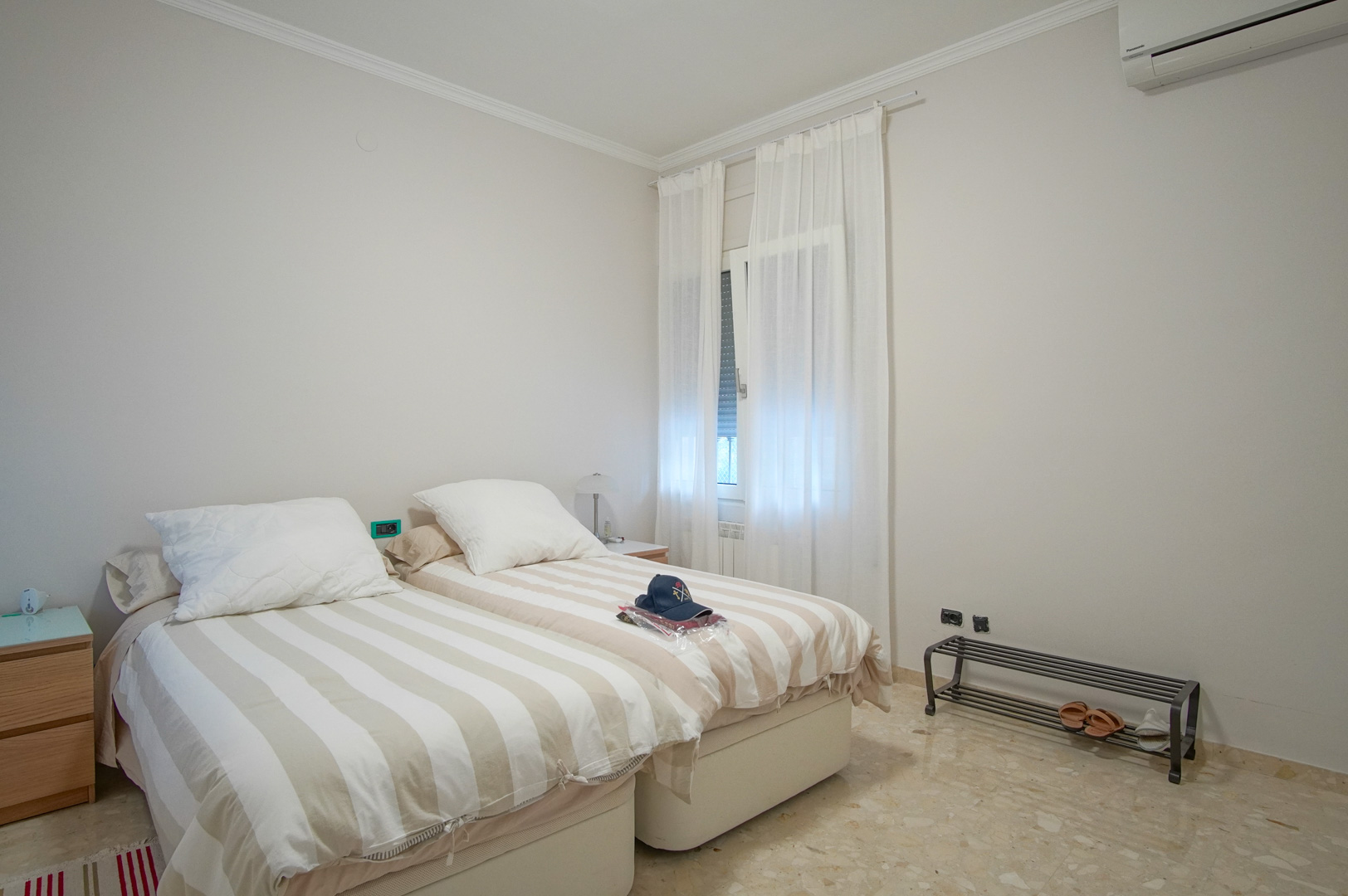 Chambre lumineuse avec deux lits simples, sol en marbre, climatisation réversible et fenêtre habillée de rideaux blancs laissant passer la lumière.