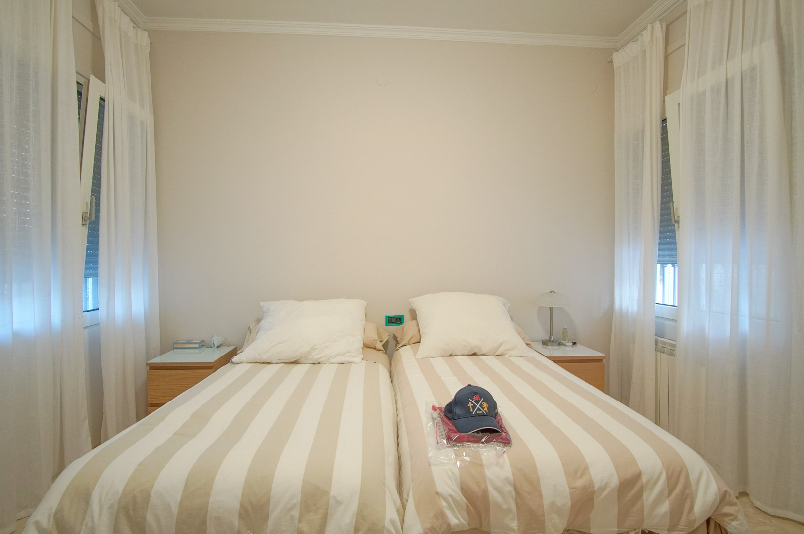 Chambre lumineuse avec deux lits simples, tables de chevet en bois et doubles fenêtres avec rideaux blancs offrant une atmosphère sereine et aérée.