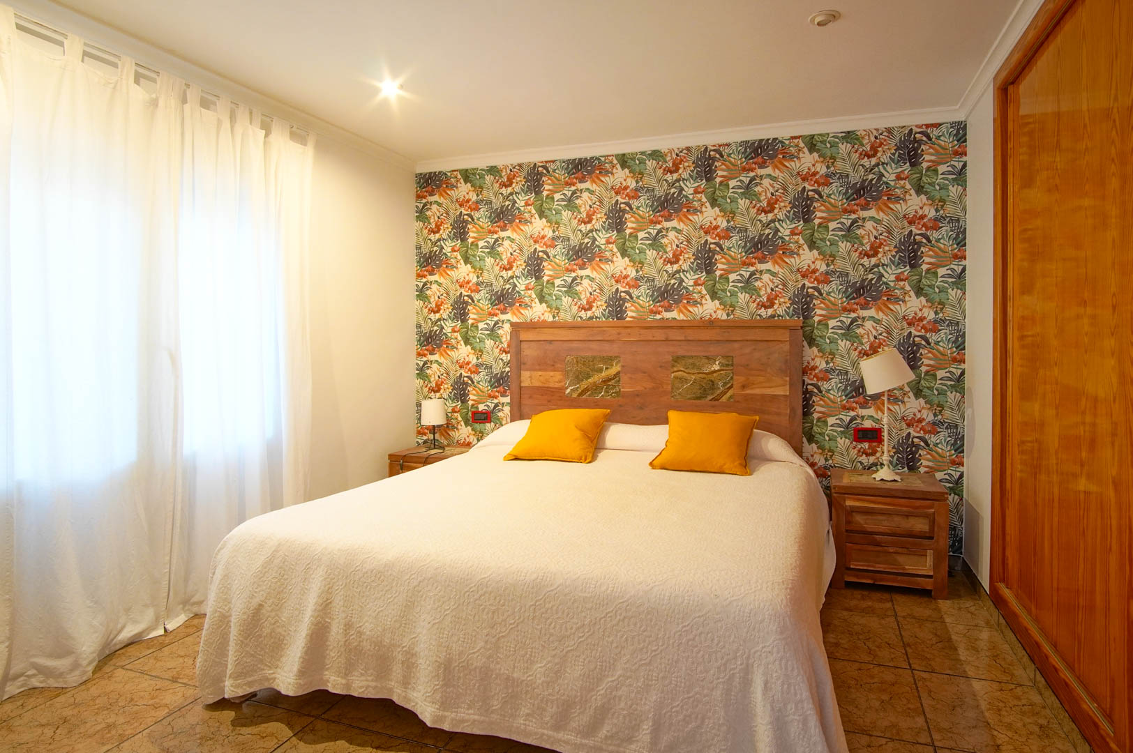 Chambre lumineuse avec papier peint tropical, tête de lit en bois avec inserts en pierre et placards intégrés en bois.