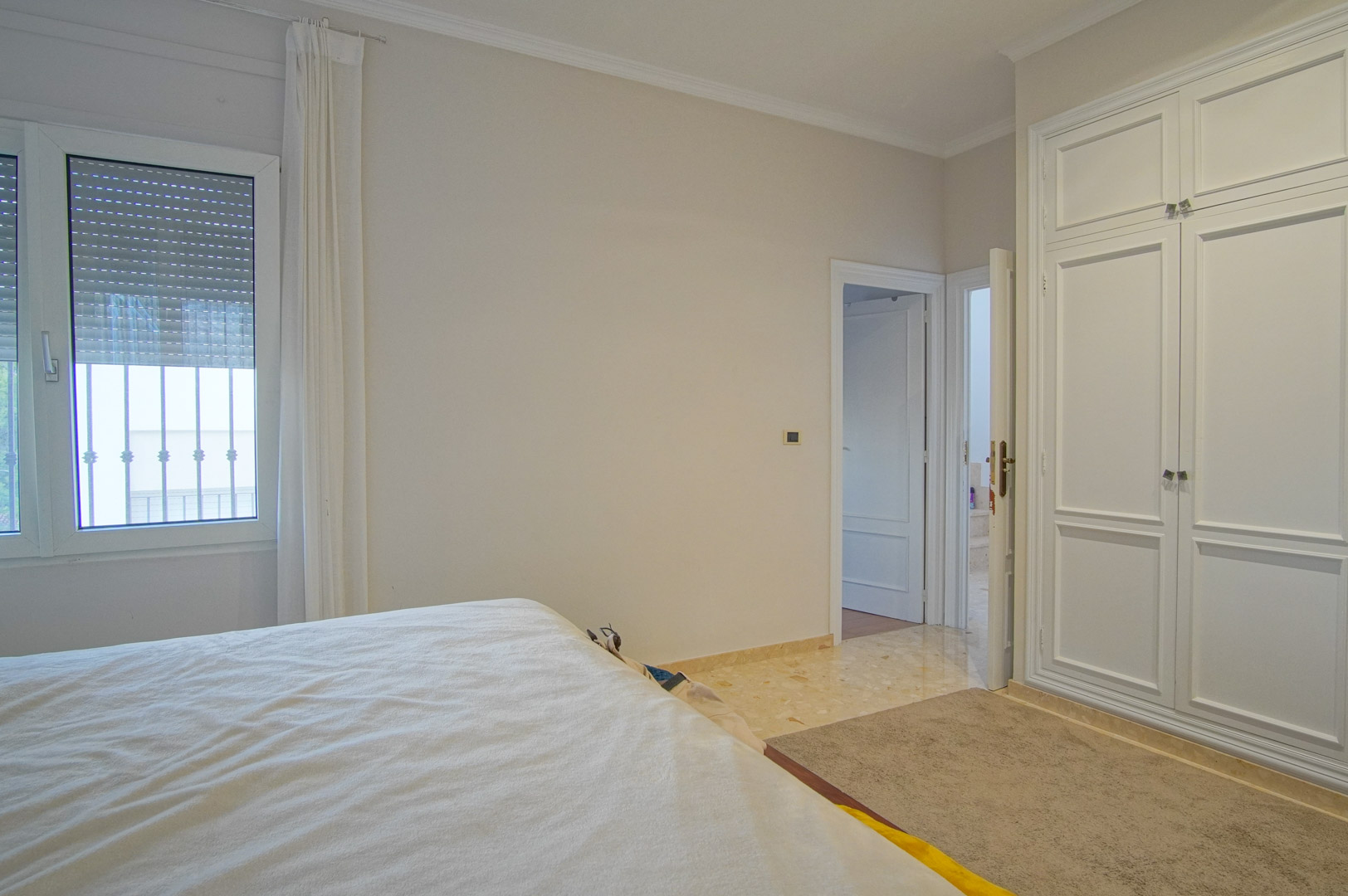 Chambre lumineuse avec placards intégrés blancs, sol en marbre et fenêtre avec volets roulants. Les tons neutres apportent une atmosphère calme et élégante.