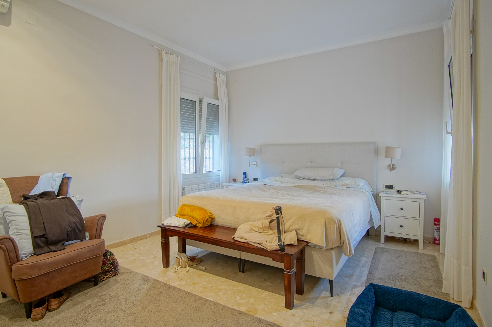 Chambre lumineuse avec sol en marbre, lit double à tête de lit rembourrée et banc en bois. Les fenêtres avec volets assurent une belle luminosité naturelle.