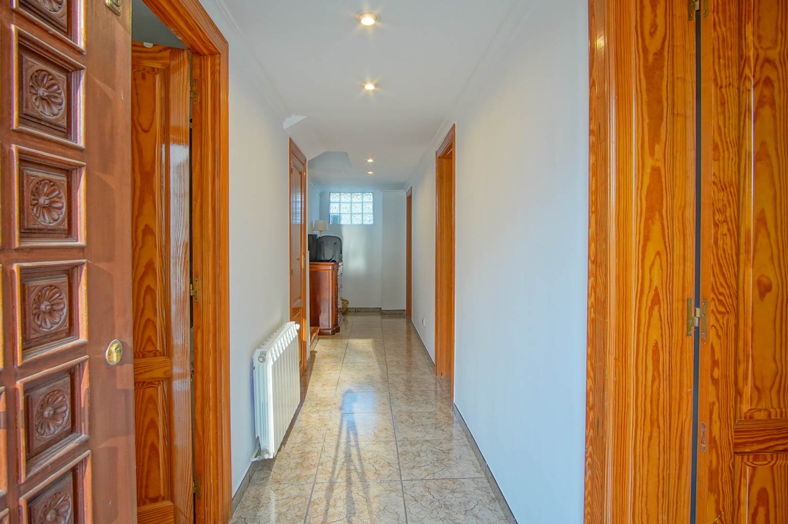 Couloir lumineux avec sol en grès cérame, portes en bois sculpté et éclairage encastré. Équipé d'un radiateur blanc et d'une fenêtre en briques de verre.