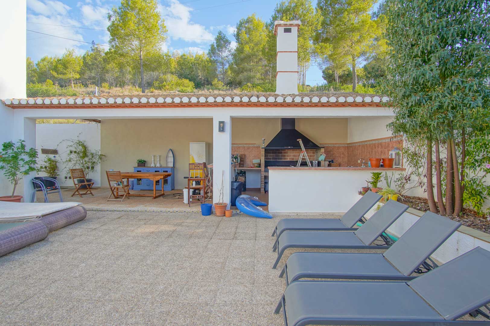 Cuisine d'été couverte avec barbecue, coin repas et terrasse en pierre avec transats, offrant une vue directe sur la pinède du parc naturel du Montgó.