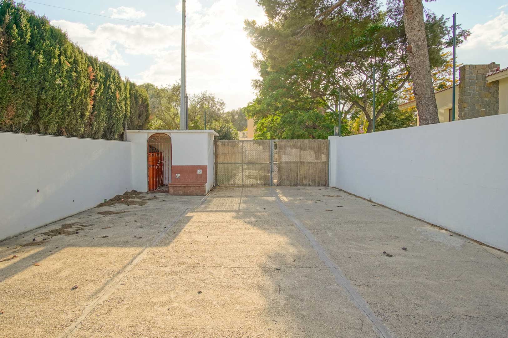 Entrée de la propriété avec une allée pavée, des murs d'enceinte blancs et un portail sécurisé, entourée de pins et de haies de cyprès.