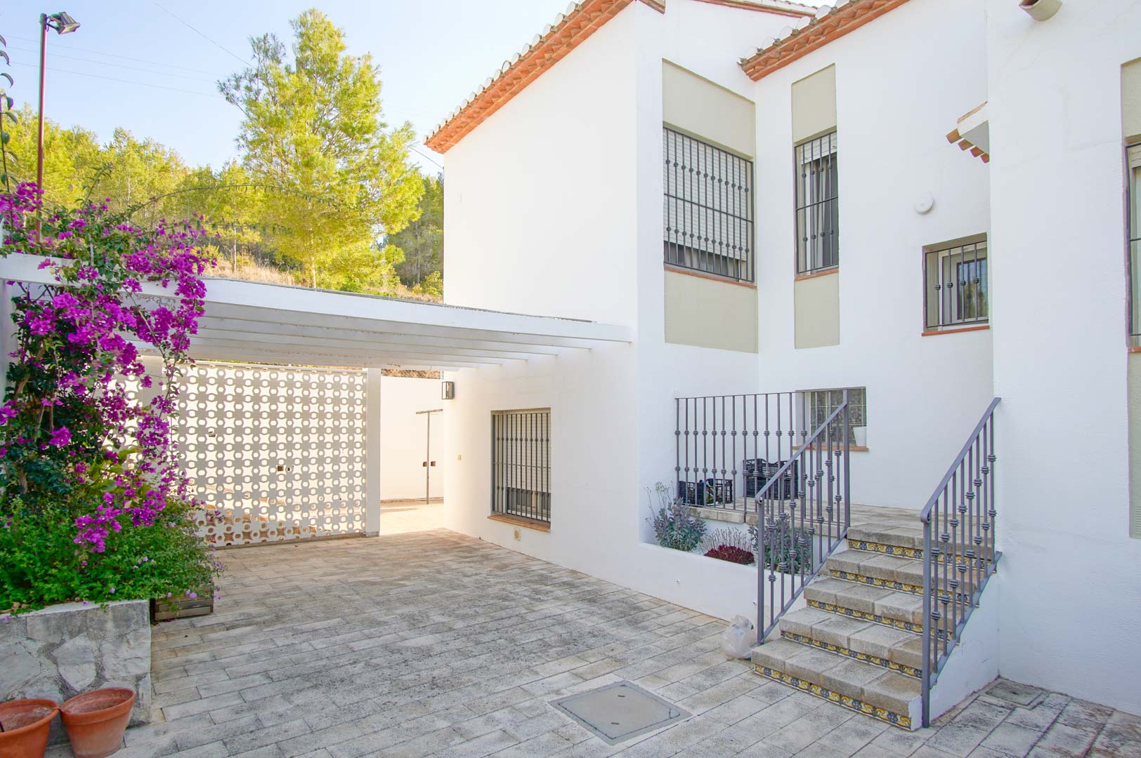 Entrée extérieure avec pavage en pierre, parking couvert par une pergola blanche et escalier d'accès orné de carreaux décoratifs et d'une rampe en fer forgé.
