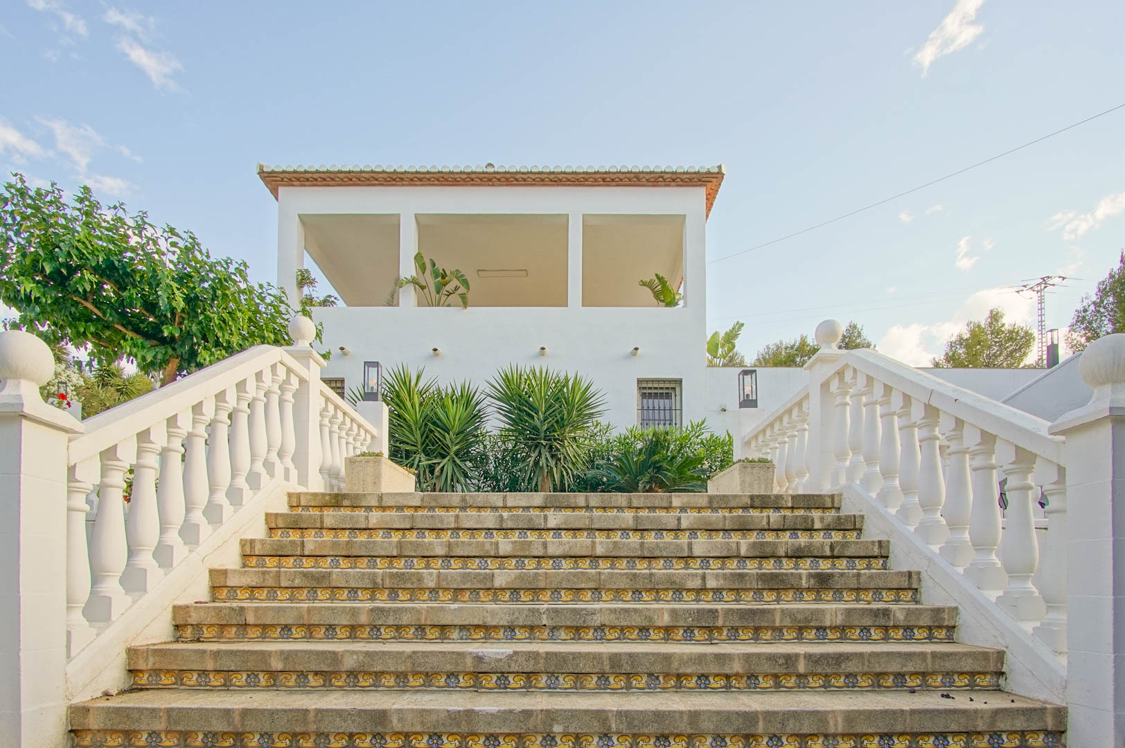 Escalier extérieur en pierre avec contremarches en céramique décorée et balustrade blanche, menant à la terrasse couverte et à la façade de la villa.