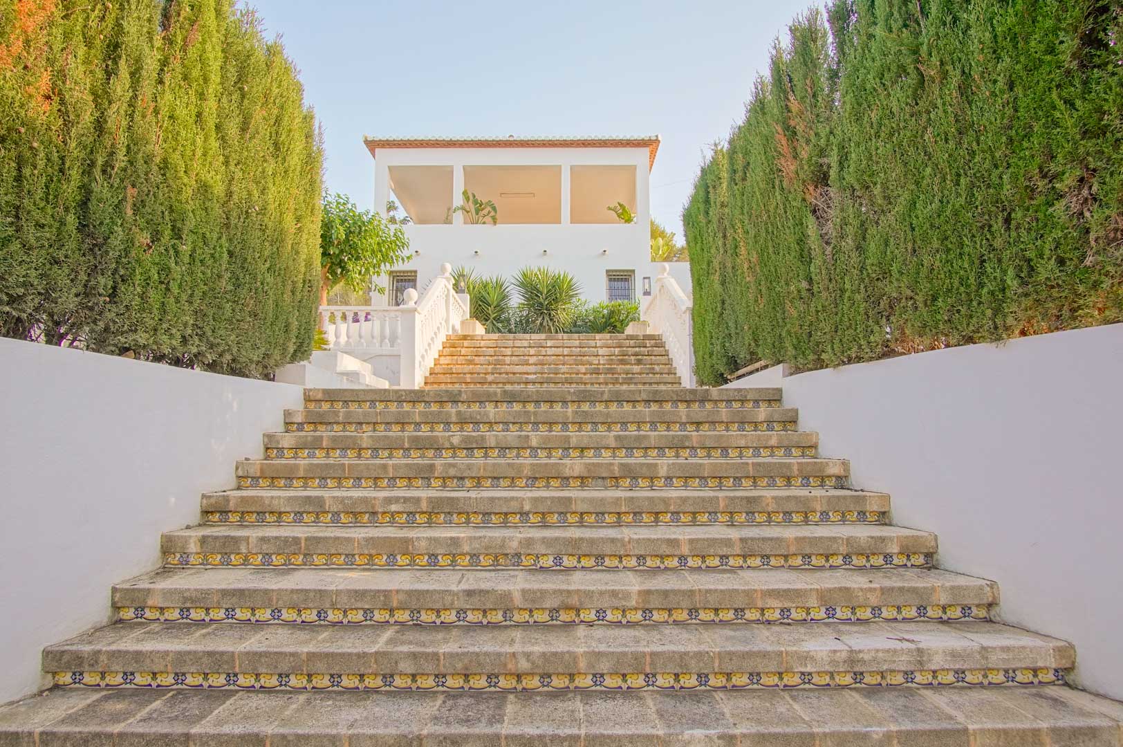 Escalier monumental en pierre avec contremarches en céramique décorative, encadré par de hautes haies de cyprès menant à la façade principale de la propriété.