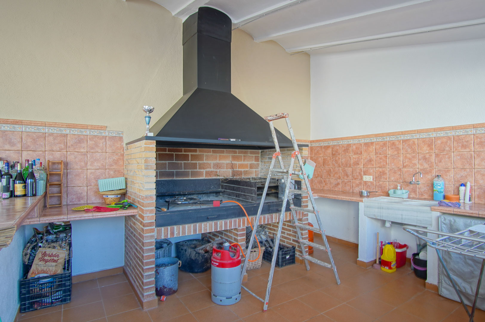 Espace barbecue maçonné avec hotte en métal noir, évier en pierre et plans de travail carrelés dans des tons terre cuite pour les repas en extérieur.