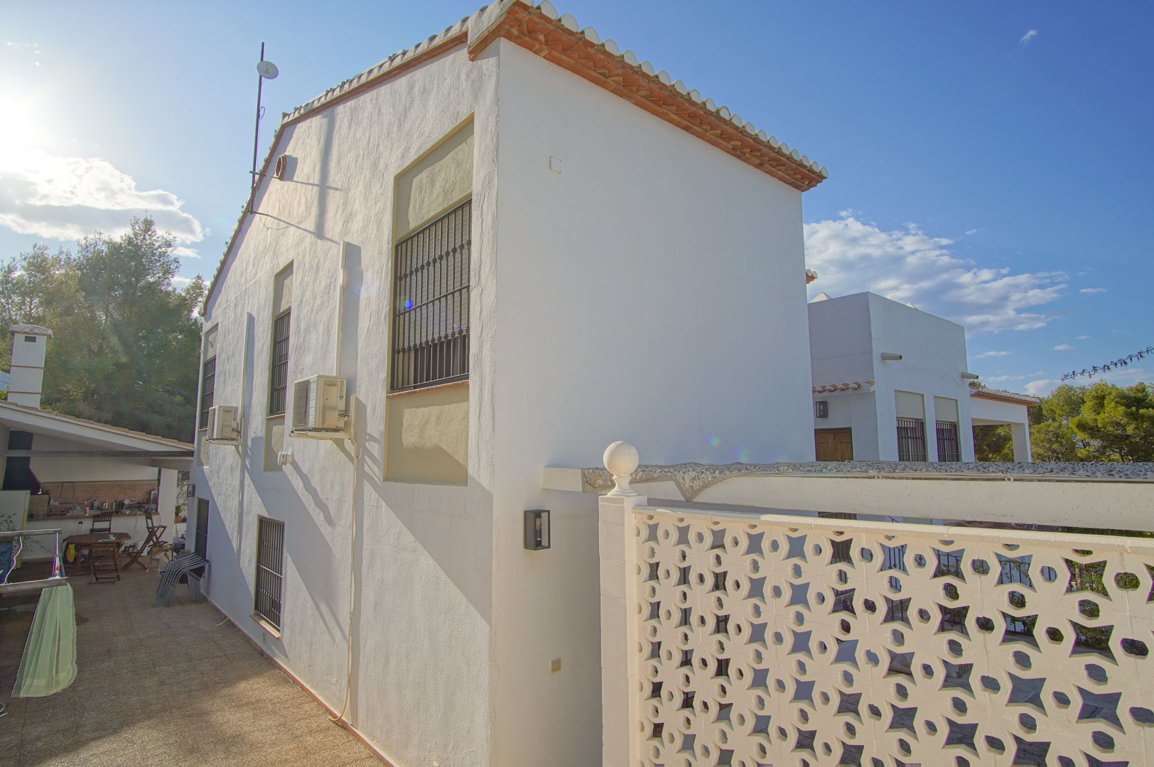 Façade latérale de la villa avec murs blancs, fenêtres sécurisées et un passage pavé menant à une cuisine d'été et un espace barbecue.