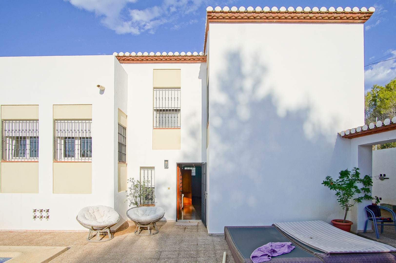 Façade méditerranéenne blanche avec fenêtres à barreaux, terrasse en pierre équipée de mobilier de jardin et accès direct vers l'intérieur de la villa.