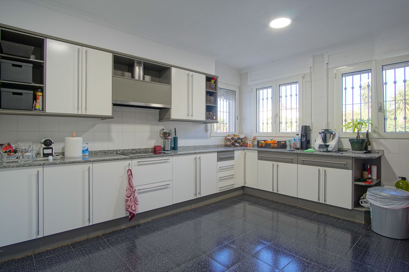 Grande cuisine indépendante avec des meubles blancs et gris, un plan de travail en granit et un sol en carrelage sombre. Plusieurs fenêtres apportent une belle luminosité.