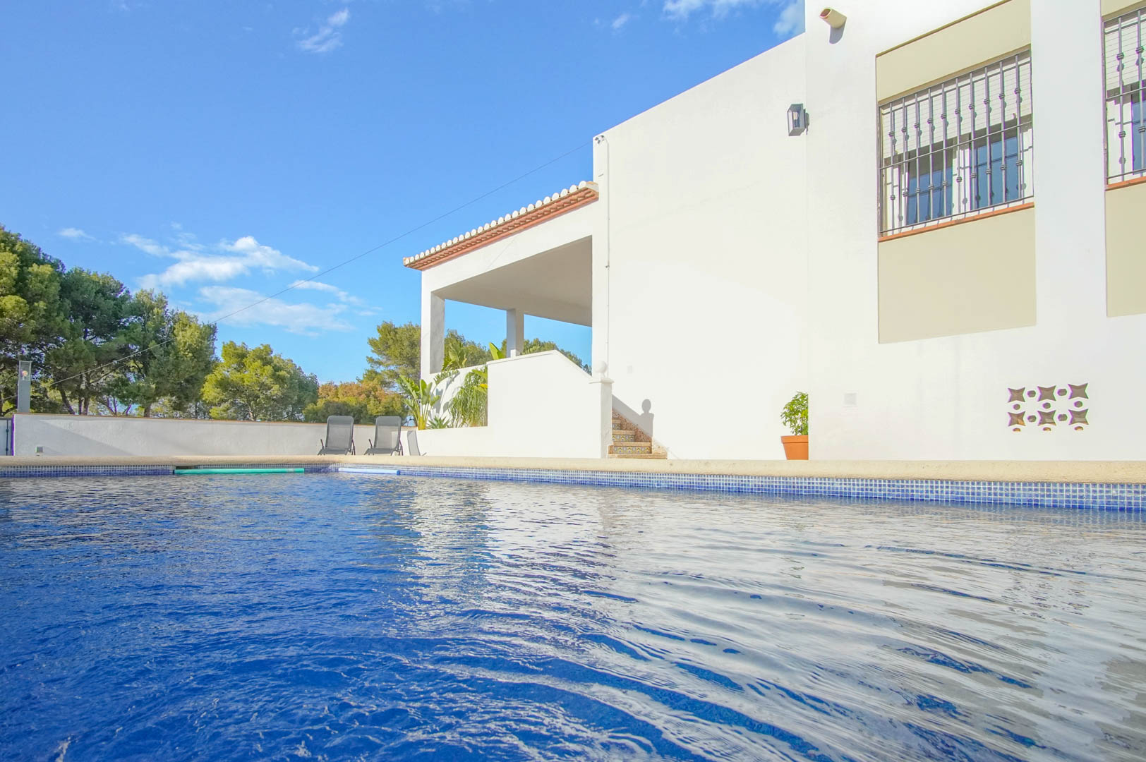 Piscine privée avec mosaïque bleue bordant la façade blanche de la villa, dotée d'une terrasse couverte, d'escaliers carrelés et entourée de pins méditerranéens.