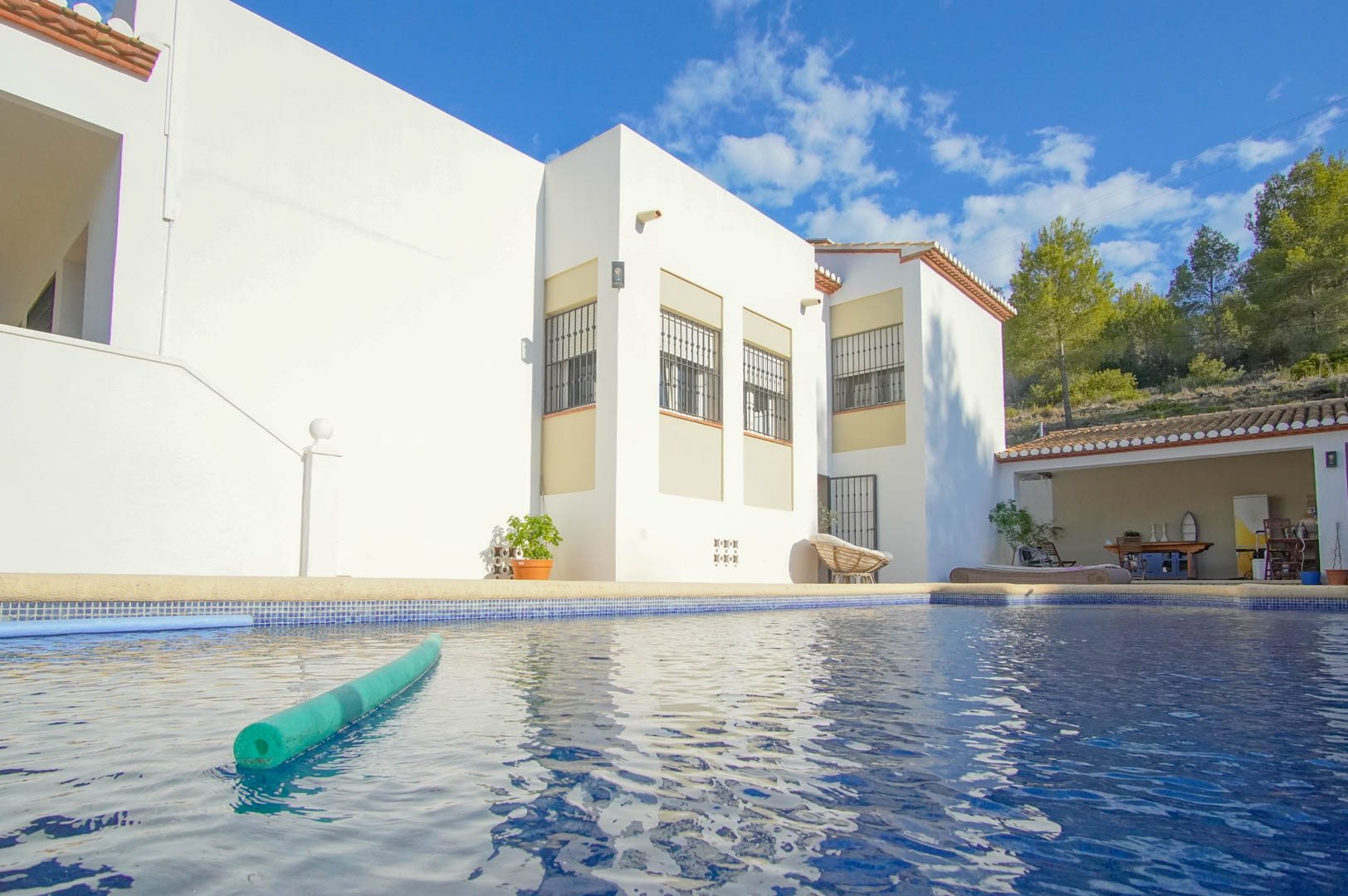 Piscine privée avec mosaïque bleue bordée par la façade blanche de la villa, comprenant une terrasse couverte et un jardin arboré en arrière-plan.