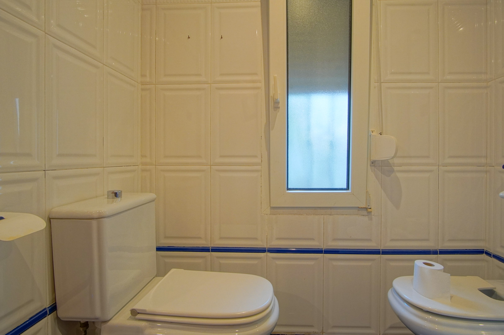 Salle de bain avec carrelage blanc en relief et liseré bleu. Équipée d'un WC, d'un bidet et d'une fenêtre en verre dépoli pour une lumière naturelle.