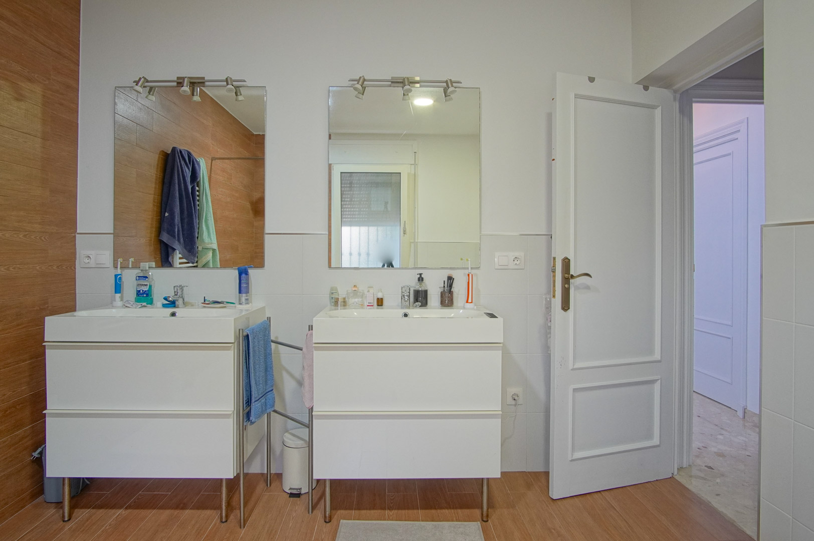 Salle de bain avec double vasque sur meubles blancs, miroirs individuels éclairés et finitions murales mêlant carrelage blanc et grès cérame aspect bois.