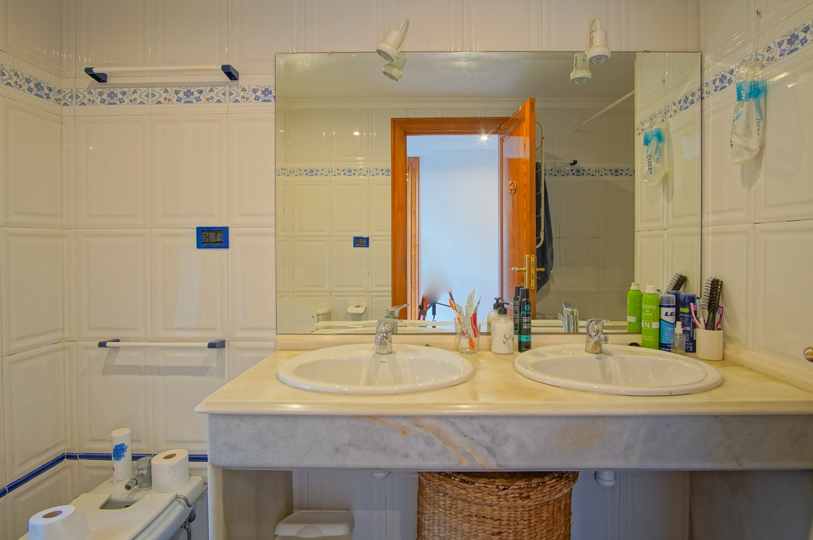 Salle de bain avec double vasque sur plan de travail en marbre, carrelage blanc avec frise florale bleue et un grand miroir éclairé.