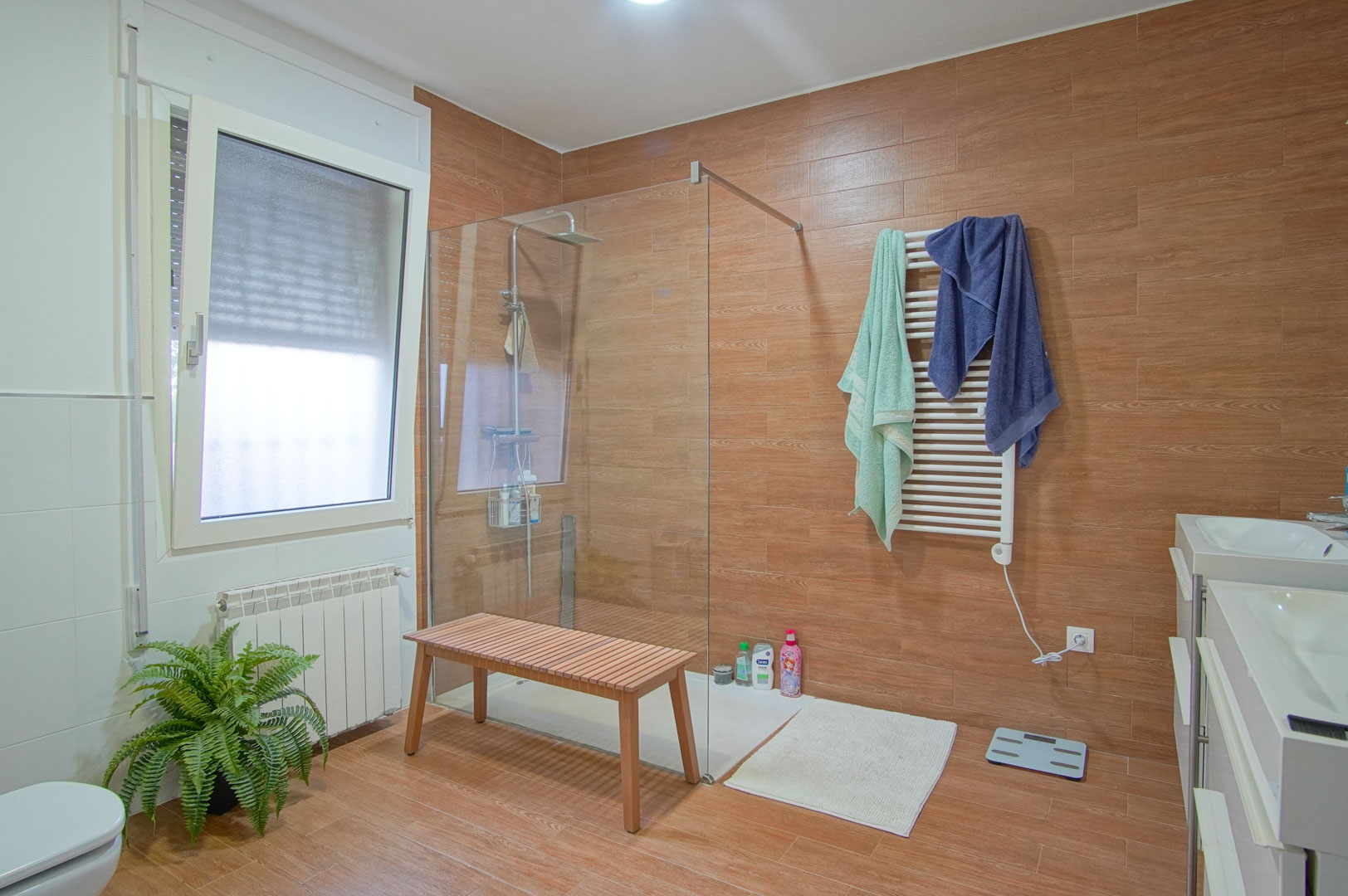 Salle de bain moderne avec carrelage effet bois, douche à l'italienne avec paroi en verre, double vasque et radiateur sèche-serviettes blanc.