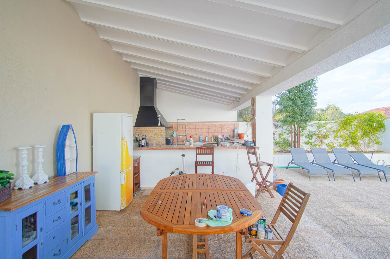 Terrasse couverte avec cuisine d'été, barbecue maçonné et plafond à poutres blanches. Comprend un coin repas en bois avec vue sur les bains de soleil et la piscine.