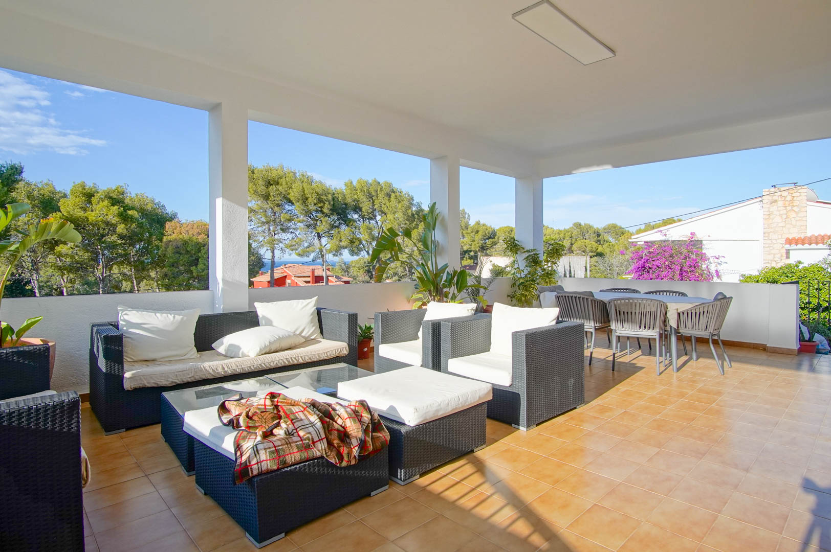 Terrasse couverte avec salon en rotin, espace repas extérieur et sol en grès, offrant une vue dégagée sur la pinède et la mer Méditerranée.