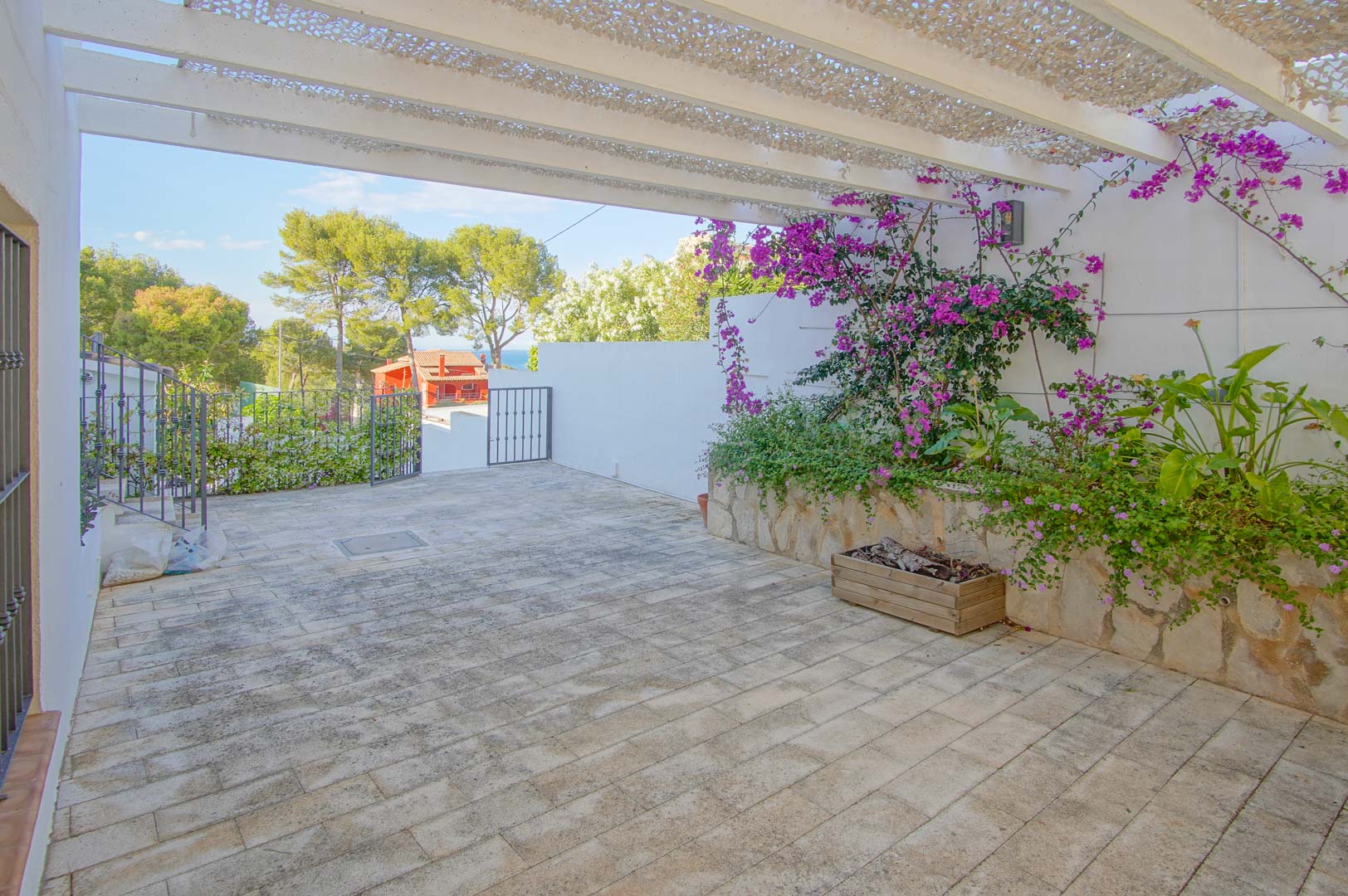 Terrasse extérieure pavée avec pergola blanche et filet d'ombrage, agrémentée de jardinières en pierre et bougainvilliers, offrant une vue dégagée sur la mer et la nature.