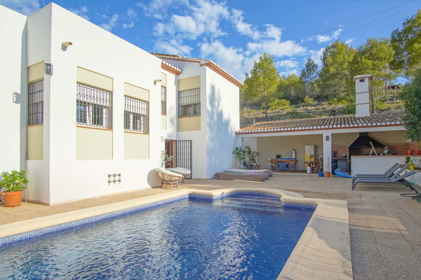 Vue extérieure avec piscine privée, terrasse spacieuse et cuisine d'été couverte avec barbecue, bordée par la forêt de pins du Montgó.