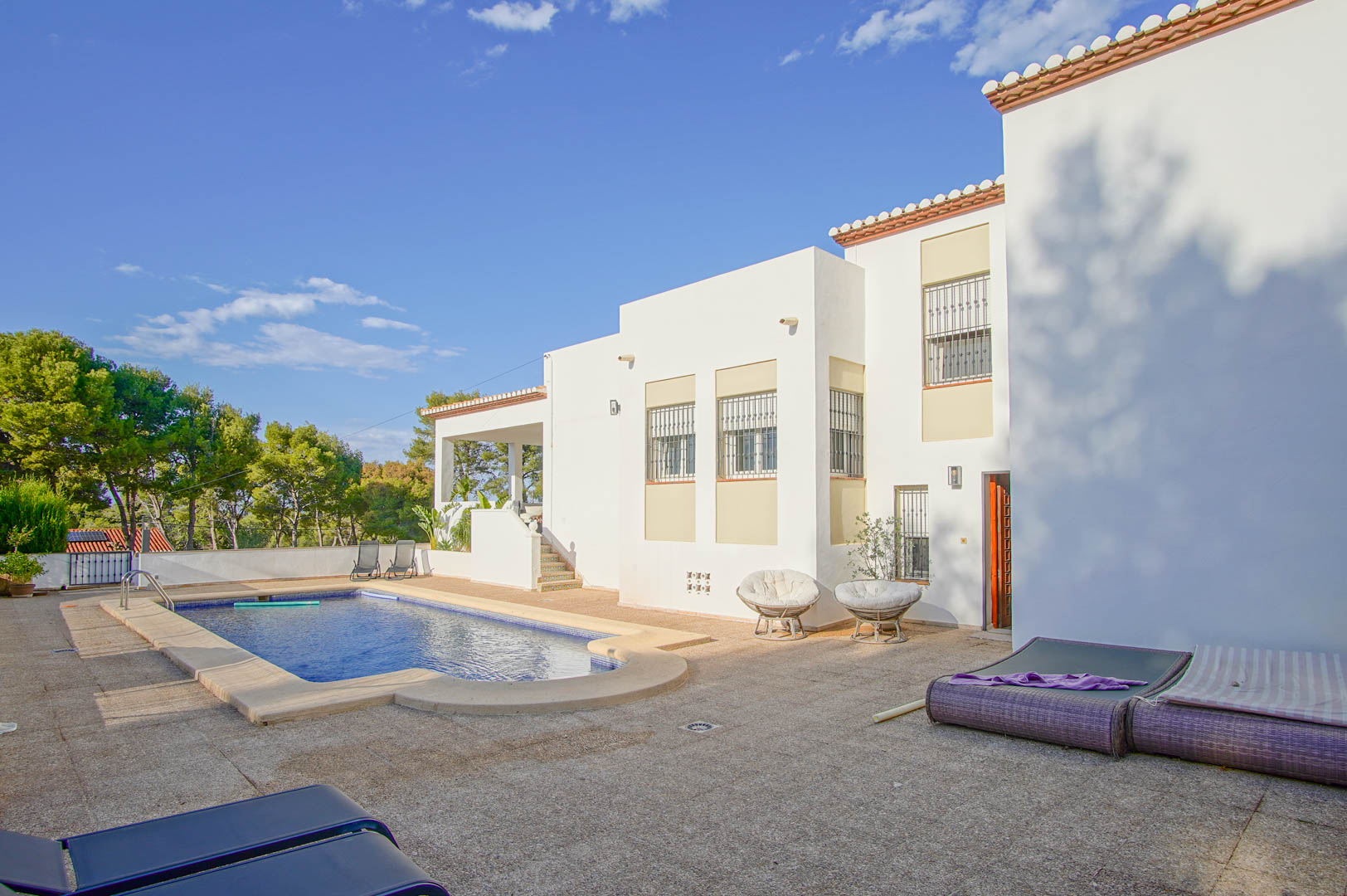 Vue extérieure de la villa à façade blanche, avec sa piscine privée et sa grande terrasse pavée entourée d'une pinède et de végétation méditerranéenne.
