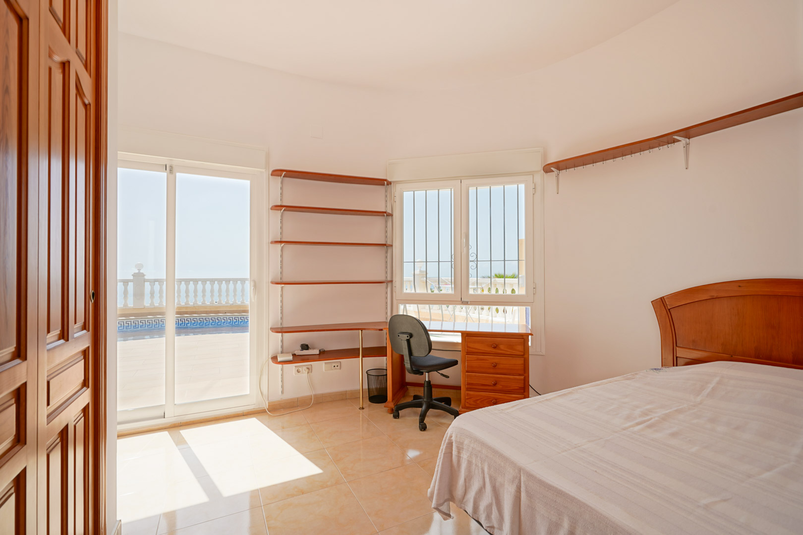 Chambre avec coin bureau, étagères en bois et accès direct à la terrasse par des portes coulissantes. Sol en grès et luminosité naturelle optimale.