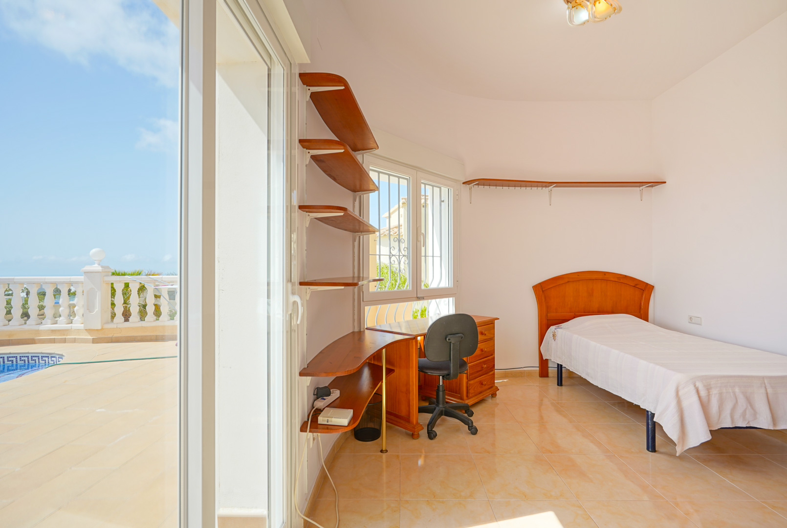 Chambre avec lit simple, bureau en bois et étagères intégrées. Les baies vitrées coulissantes offrent un accès direct à la terrasse de la piscine.