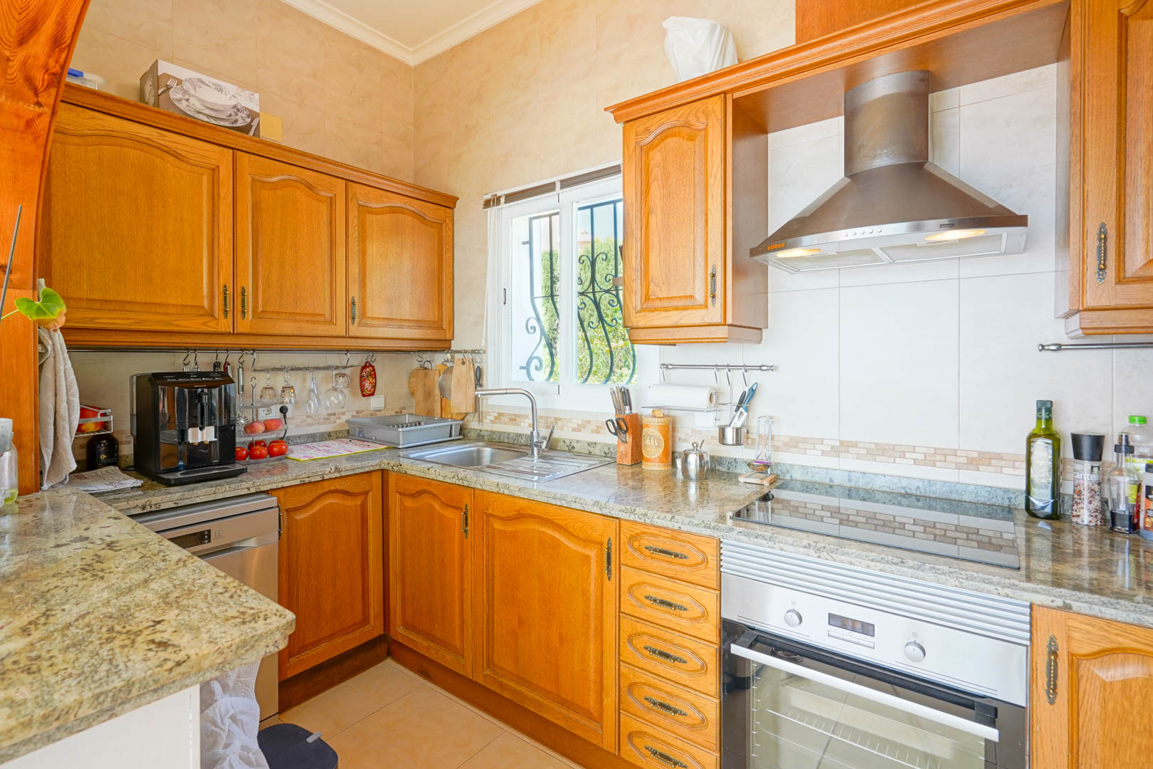 Cuisine semi-ouverte avec meubles en bois classique, plans de travail en granit et électroménagers en inox, comprenant une hotte aspirante et un four encastré.