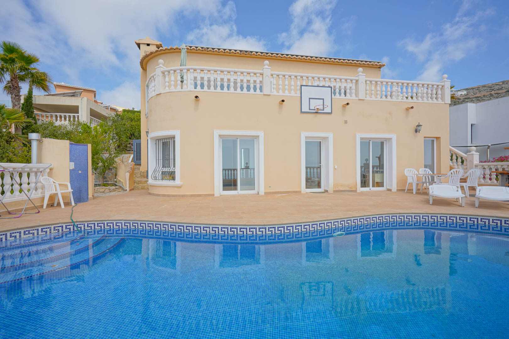 Façade incurvée de la villa avec piscine en mosaïque bleue, terrasse en terre cuite, balustrades blanches et coin salon extérieur sous le soleil.