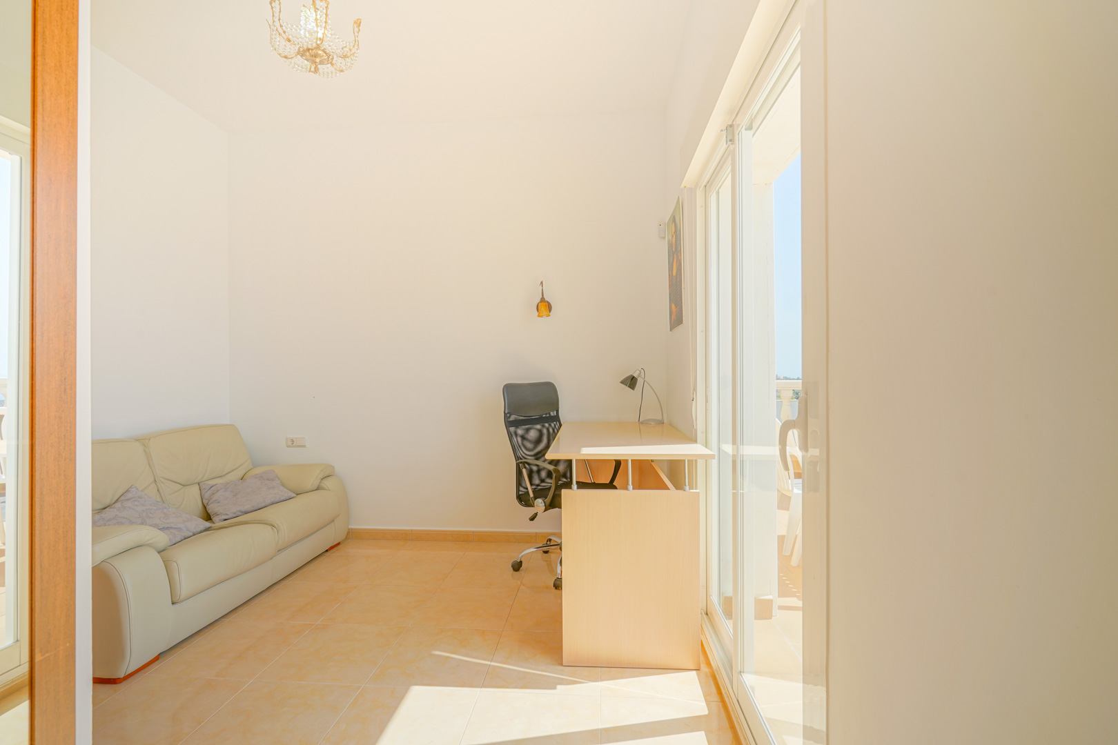Pièce lumineuse avec coin bureau, canapé en cuir beige et accès direct à la terrasse par de grandes baies vitrées. Sol en grès aux tons chauds et éclairage naturel.