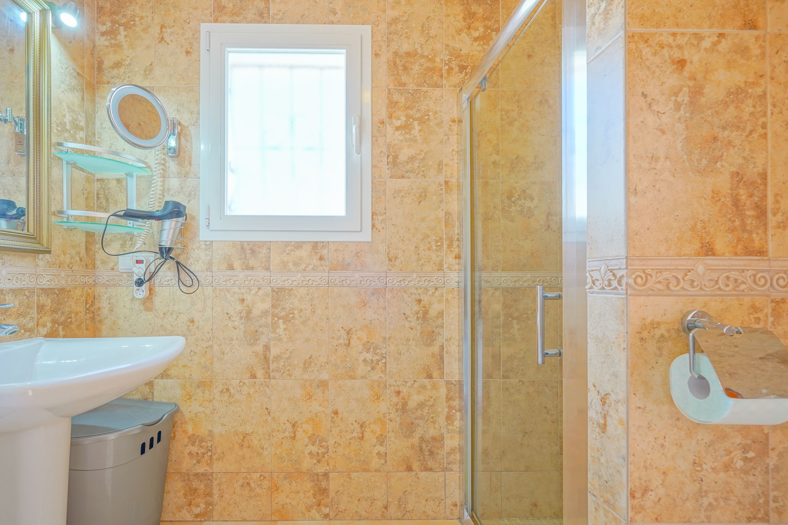 Salle de bain avec carrelage dans les tons ocre, lavabo sur colonne blanc et douche avec paroi vitrée. Une fenêtre oscillo-battante apporte de la lumière naturelle.