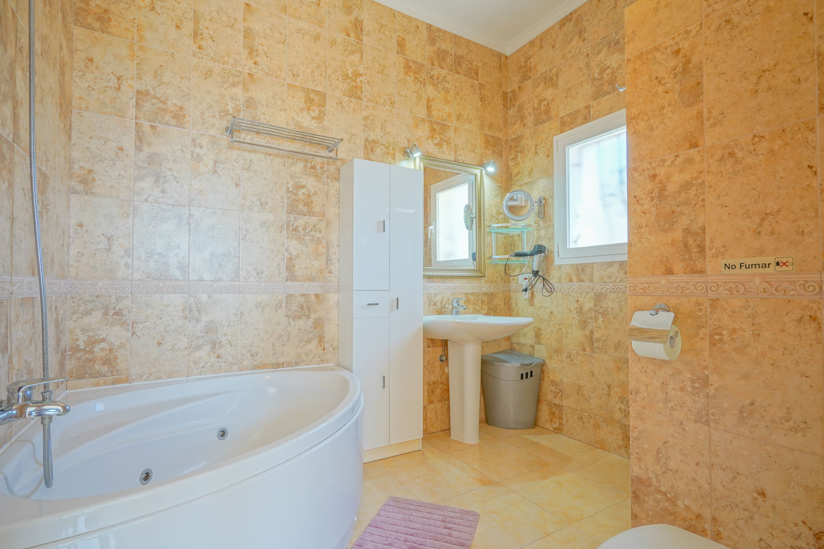 Salle de bains avec baignoire balnéo d'angle, murs revêtus de carreaux céramiques aux tons ocres et meuble de rangement blanc.