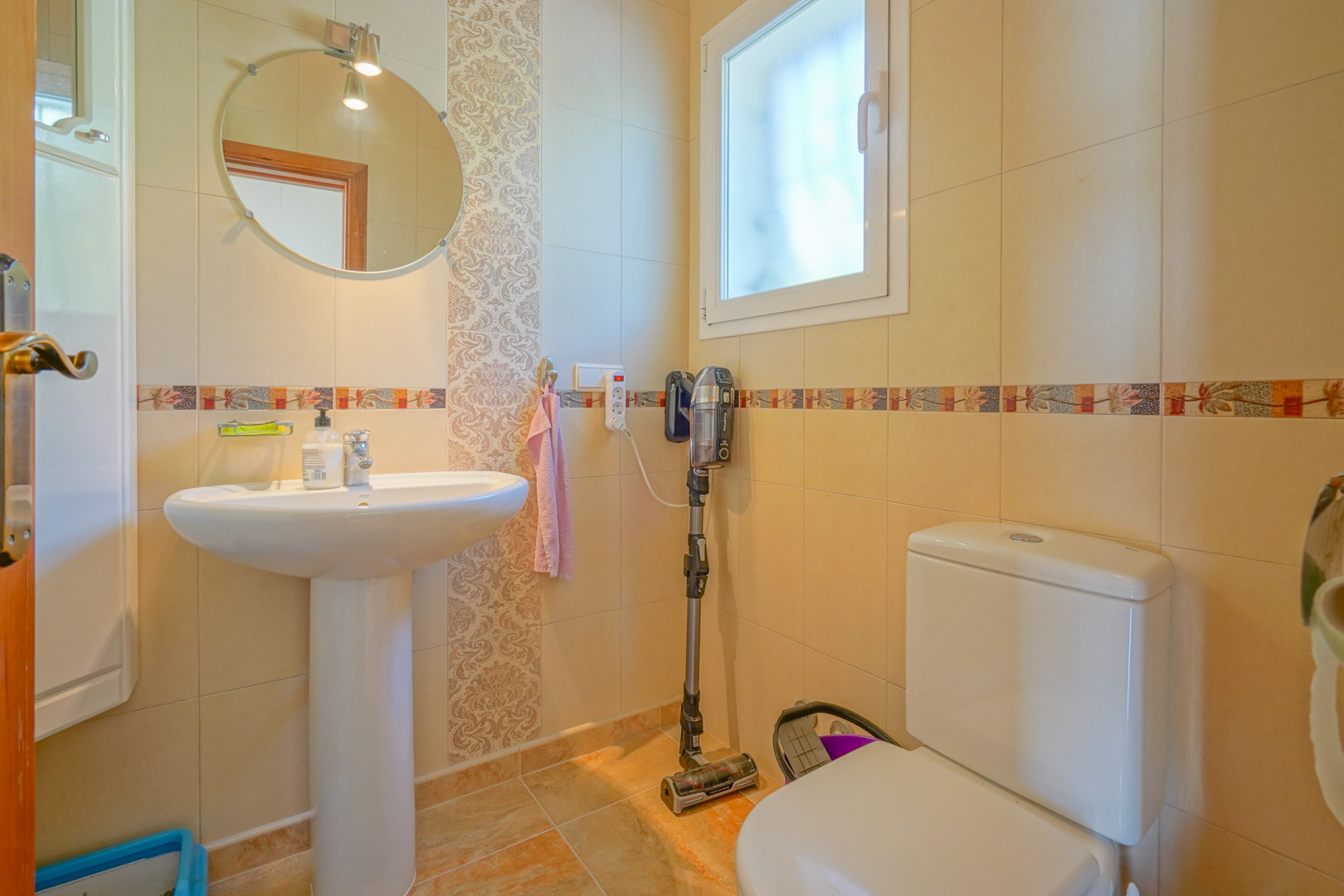 Salle de bains fonctionnelle avec lavabo sur colonne, miroir circulaire et murs carrelés aux tons crème avec frises décoratives.