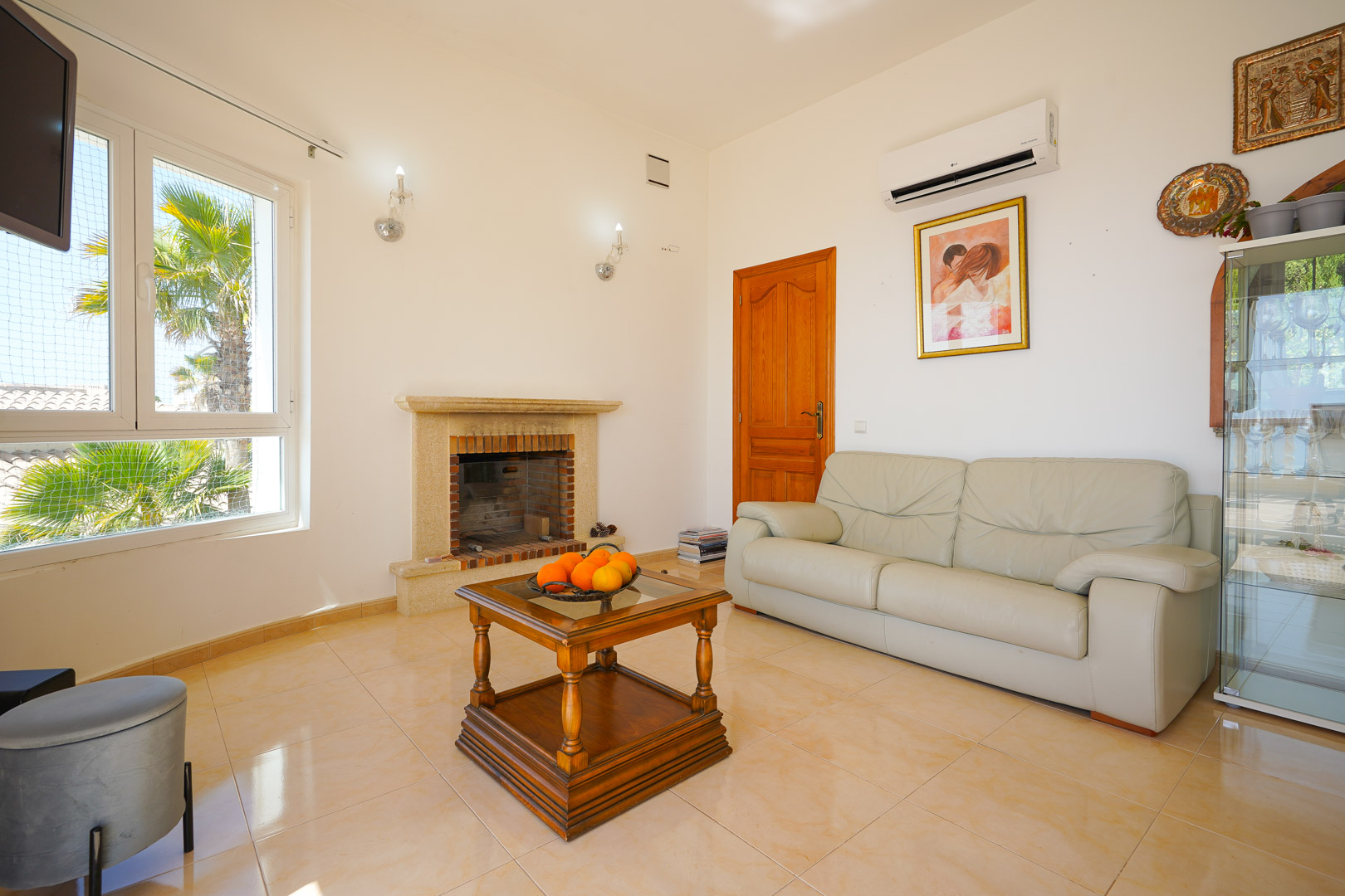 Salon avec cheminée en pierre, canapé en cuir beige et table basse en bois. La pièce dispose de la climatisation et d'une vue sur les palmiers par la fenêtre.