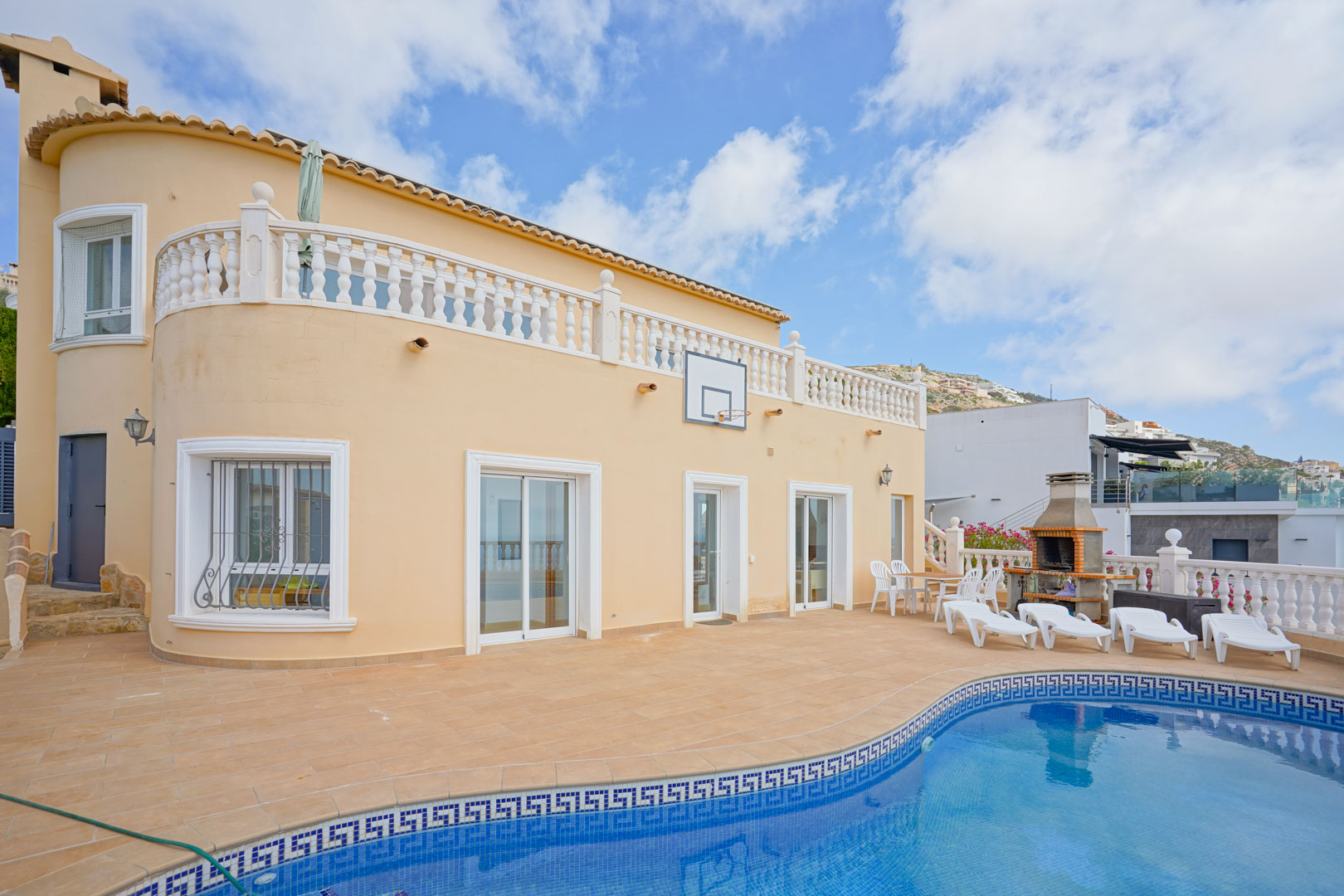 Terrasse extérieure avec piscine en mosaïque bleue, barbecue en brique et espace solarium. La façade incurvée présente des fenêtres blanches et une balustrade classique à l'étage.